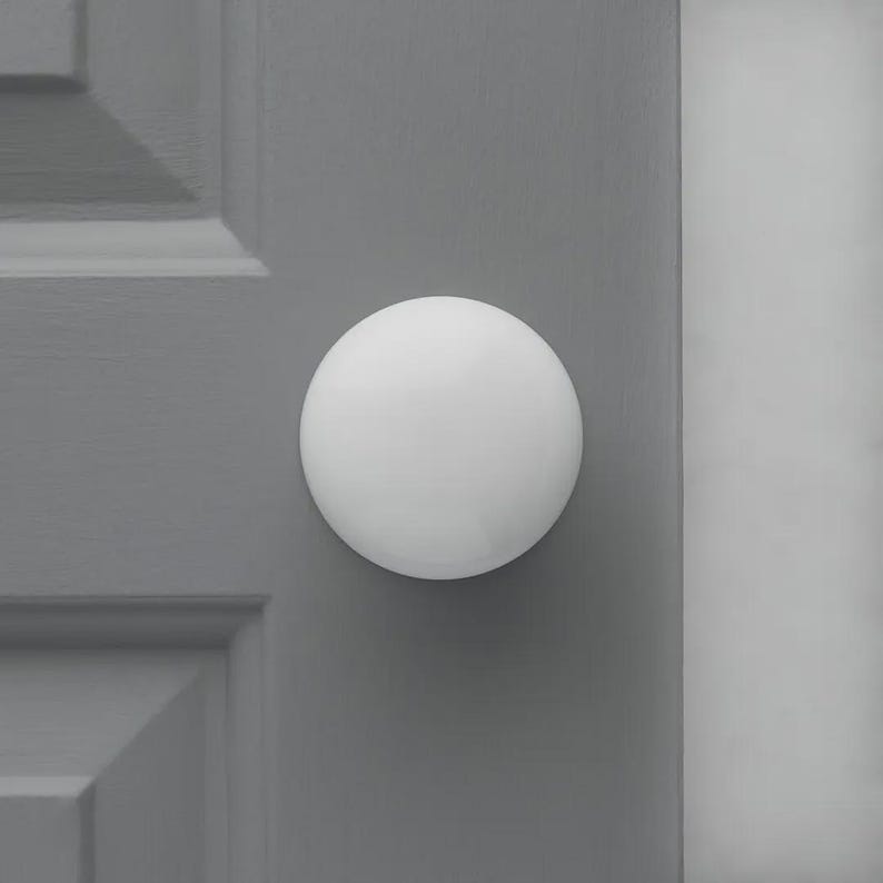 Op de afbeelding: Een witte, bolvormige deurknop is gemonteerd op een grijze deur. De deur heeft een paneelontwerp. De knop is gecentreerd op de deur en lijkt gemaakt van een glad, massief materiaal. De achtergrond is effen grijs.
