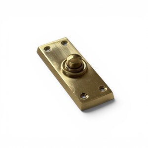 Op de afbeelding: Een rechthoekige messing deurbelknop met een ronde knop in het midden. De deurbelknop heeft een geborstelde afwerking en vier schroefgaten. De knop heeft een warme goudkleur en staat tegen een witte achtergrond.