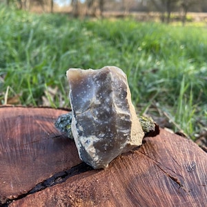 May include: A gray flint stone with a rough, irregular surface. The stone is sitting on a brown tree stump in a grassy field.