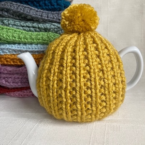 May include: A yellow knitted teapot cosy with a pom pom on top. The cosy is made with a chunky yarn and has a ribbed texture.