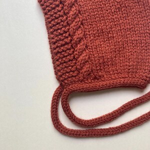 Puede incluir: Primer plano de un gorro tejido de color &oacute;xido con un dise&ntilde;o de punto de cable en el lateral. El gorro tiene dos lazos largos tejidos. El fondo es una superficie blanca lisa. El gorro est&aacute; hecho de un hilo suave y texturizado.