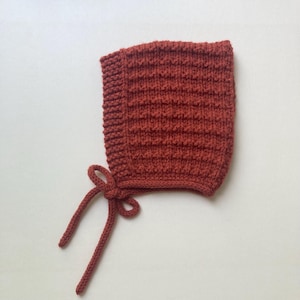 May include: A terracotta-coloured knitted baby bonnet with a textured pattern. The bonnet has a rounded shape and ties with two long, knitted strings. The background is a plain white surface.