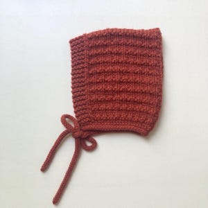 May include: A rust-coloured knitted bonnet featuring a textured horizontal ribbed pattern across the main body and a smooth garter stitch border along the face opening. The bonnet includes two matching knitted ties attached at the chin area.