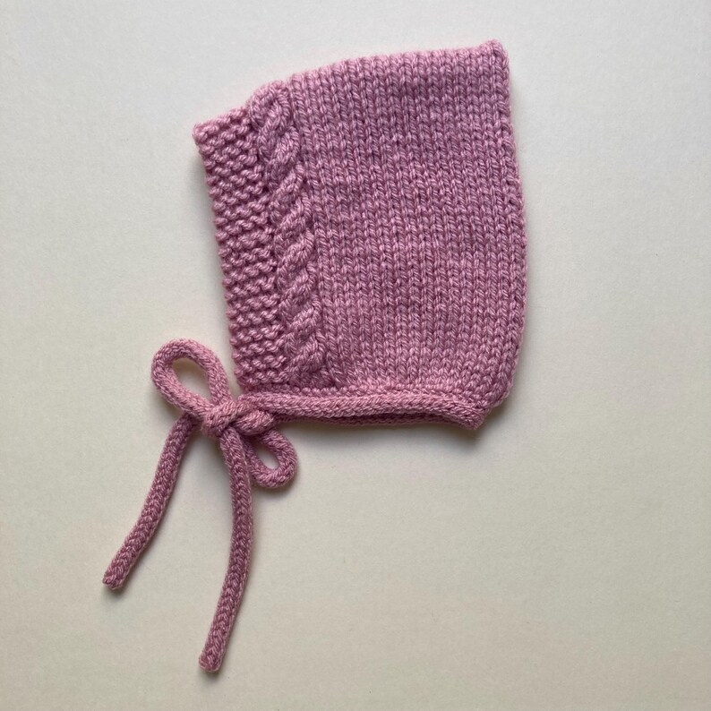 Puede incluir: Un gorro de beb&eacute; tejido en color malva suave. El gorro presenta un dise&ntilde;o de punto trenzado en el lateral y se ata con un lazo bajo la barbilla. El gorro est&aacute; hecho de un hilo suave y texturizado.