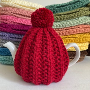 May include: A vibrant red knitted tea cosy with a pom-pom, covering a white teapot. The cosy features a textured, ribbed pattern. A stack of colourful knitted tea cosies in various colours are in the background.