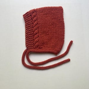 Puede incluir: Un gorro de beb&eacute; tejido de color &oacute;xido con un dise&ntilde;o de trenza en el lateral. El gorro tiene lazos largos a juego para sujetarlo bajo la barbilla. La textura de punto es visible en todo el gorro.