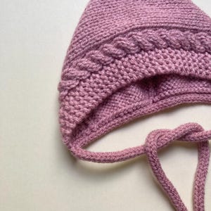 Puede incluir: Primer plano de un gorro de punto en color rosa empolvado. El gorro presenta un dise&ntilde;o de punto trenzado en la frente y un cierre de lazo. El tejido de punto suave y texturizado es adecuado para beb&eacute;s y ni&ntilde;os.