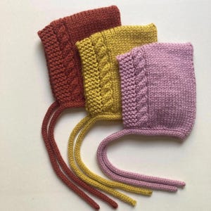May include: Three knitted baby bonnets in rust, mustard yellow, and mauve. Each bonnet has a cable knit design on the front and long ties. The bonnets are made of soft yarn. Suitable for newborns, these bonnets are a charming accessory.
