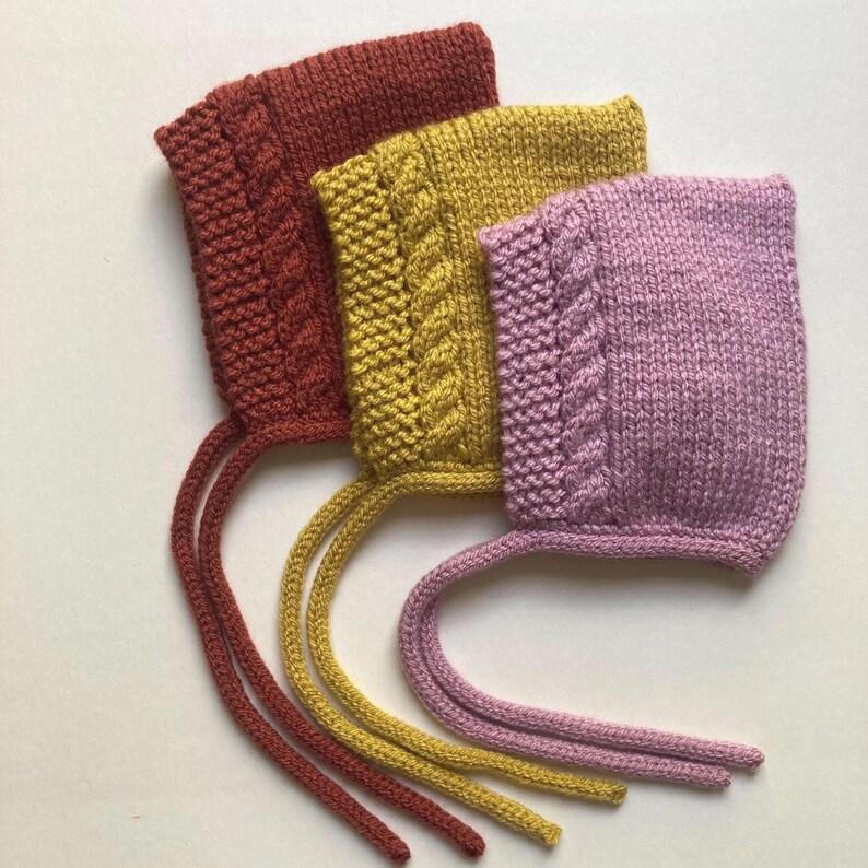 Puede incluir: Tres gorros de beb&eacute; tejidos en tonos &oacute;xido, amarillo mostaza y malva. Cada gorro presenta un dise&ntilde;o de punto trenzado en la parte delantera y lazos largos. Los gorros est&aacute;n hechos de hilo suave y son adecuados para reci&eacute;n nacidos.
