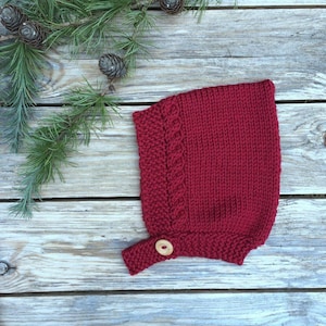 May include: A deep red knitted bonnet featuring a textured cable knit pattern along the front edge and a single light-coloured wooden button closure at the chin strap. The bonnet is constructed with a combination of garter and stockinette stitches.