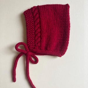 May include: A knitted red baby bonnet. The bonnet features a cable knit design on the side and ties under the chin with matching red yarn. The bonnet is made of a soft, textured yarn. The bonnet is a deep shade of red.