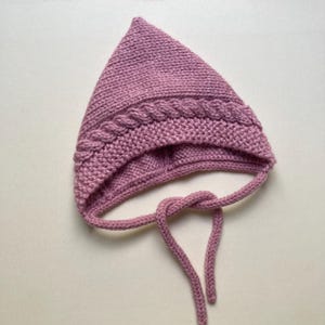 Puede incluir: Un gorro de beb&eacute; tejido, de color malva, con una parte superior puntiaguda y un dise&ntilde;o trenzado de punto de cable alrededor de la frente. El gorro tiene lazos adjuntos para asegurar debajo de la barbilla. El fondo es de color neutro s&oacute;lido.