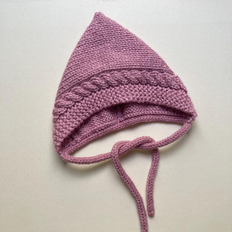Puede incluir: Un gorro de beb&eacute; tejido, de color malva, con una parte superior puntiaguda y un dise&ntilde;o trenzado de punto de cable alrededor de la frente. El gorro tiene lazos adjuntos para asegurar debajo de la barbilla. El fondo es de color neutro s&oacute;lido.