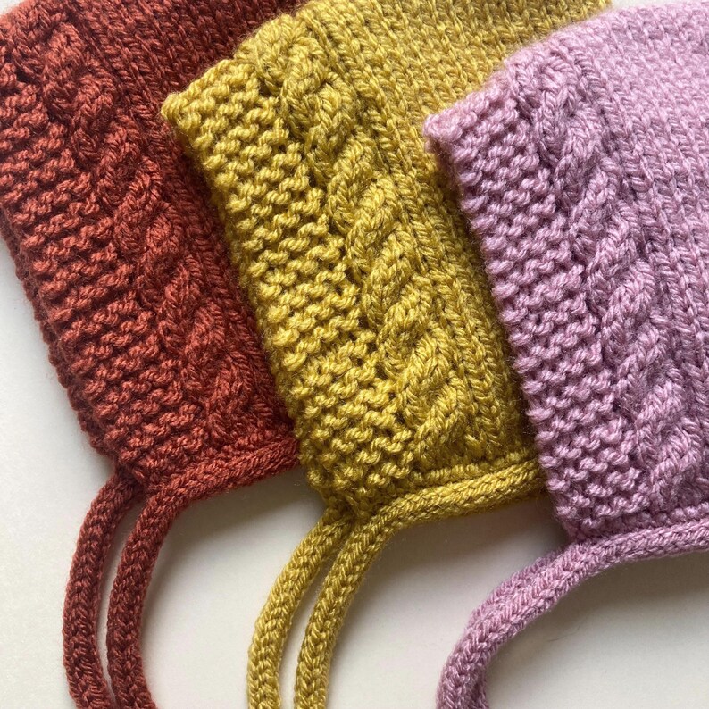 Puede incluir: Primer plano de tres gorros de beb&eacute; tejidos en tonos &oacute;xido, amarillo mostaza y lavanda. Cada gorro presenta un dise&ntilde;o de punto de cable y lazos adjuntos. Los gorros est&aacute;n hechos de hilo suave y son adecuados para beb&eacute;s.