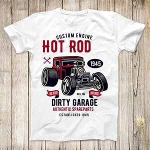 Puede incluir: Camiseta blanca con una ilustración roja, negra y blanca de un coche de carreras vintage. El texto "CUSTOM ENGINE HOT ROD" está por encima del coche, y el texto "DIRTY GARAGE AUTHENTIC SPAREPARTS ESTABLISHED 1945" está por debajo del coche.