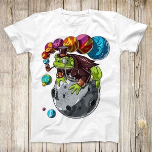 May include: A white t-shirt with a green frog wearing a top hat and monocle sitting on a grey moon. The frog is surrounded by colorful planets.