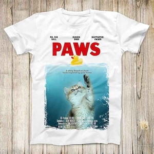 May include: White t-shirt with a graphic of a movie poster for a film called "PAWS". The poster features a cat reaching for a yellow rubber ducky in a blue pool. The text on the poster reads "A story based on facts reals inspired by your home".