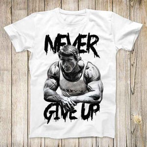Puede incluir: Camiseta blanca con una ilustración en blanco y negro de un hombre musculoso con el texto "NEVER GIVE UP" en una fuente que gotea.