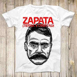 May include: White t-shirt with a black and white illustration of Emiliano Zapata, a Mexican revolutionary leader. The text "ZAPATA! ¡TIERRA Y LIBERTAD!" is printed in red on the shirt.