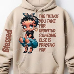 Op de afbeelding: Een beige hoodie met een cartoon afbeelding van een vrouw met een blauwe bandana en gescheurde jeans. De tekst "The things you take for granted someone else is praying for" is in bruin op de hoodie gedrukt.