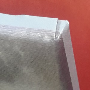 May include: Close-up of a corner of a light gray object with a textured surface. The object has a white border and is set against a red background. The corner is sharply angled.