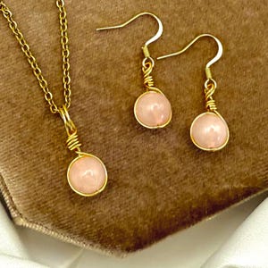 May include: A gold-tone jewellery set comprising a necklace and earrings. Each piece features a round, pale pink gemstone encased in gold wire. The necklace has a fine chain, and the earrings have hook fastenings. The set is presented on a brown surface.