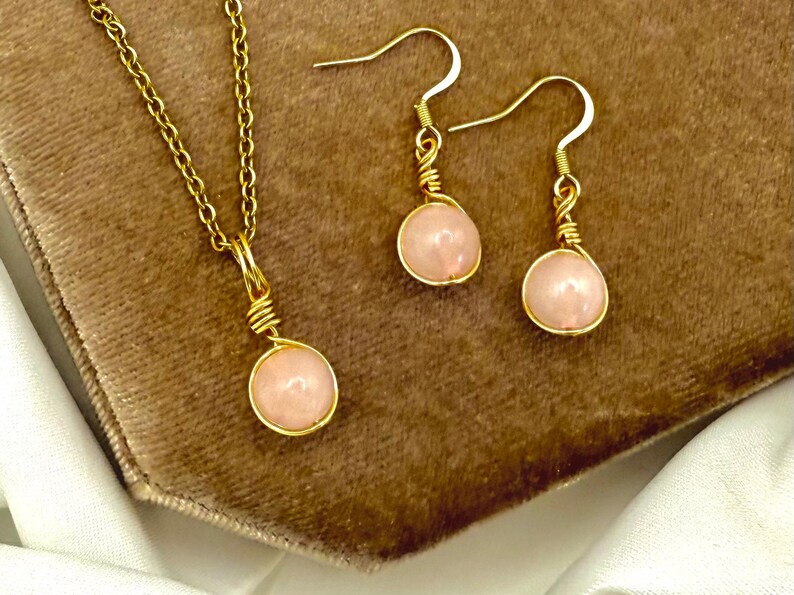 May include: A gold-tone jewellery set comprising a necklace and earrings. Each piece features a round, pale pink gemstone encased in gold wire. The necklace has a fine chain, and the earrings have hook fastenings. The set is presented on a brown surface.