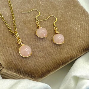 May include: A gold-tone necklace and earring set with rose quartz beads. The necklace has a fine chain and a single rose quartz pendant. The earrings have a hook design with matching rose quartz beads. The jewellery is displayed on a brown surface.