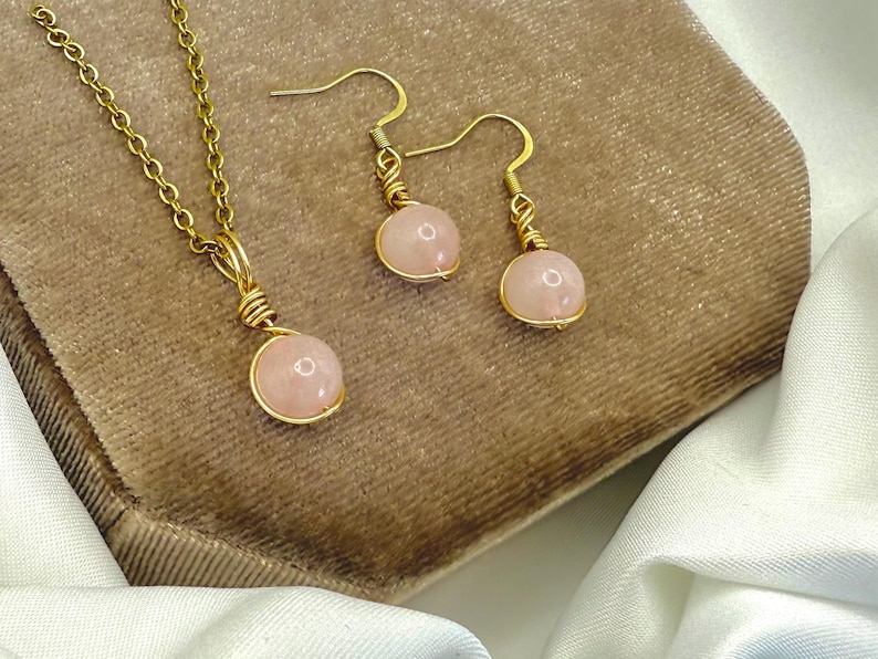 May include: A gold-tone necklace and earring set with rose quartz beads. The necklace has a fine chain and a single rose quartz pendant. The earrings have a hook design with matching rose quartz beads. The jewellery is displayed on a brown surface.