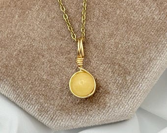 Puede incluir: Un collar dorado con un pequeño colgante redondo amarillo. El colgante está envuelto en alambre y cuelga de una delicada cadena. El collar se exhibe sobre una superficie texturizada de color beige.