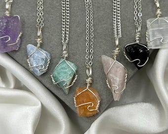 May include: A collection of gemstone pendants on silver chains. The pendants feature various raw crystals, including amethyst, blue calcite, green aventurine, orange calcite, rose quartz, black obsidian, and selenite. Each stone is wrapped in silver wire.