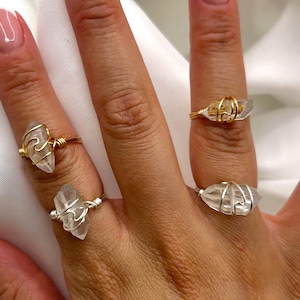 Puede incluir: Cuatro anillos con piedras de cristal transparente se muestran en los dedos. Dos anillos tienen engastes de alambre dorado y dos tienen engastes de alambre plateado. Los anillos están en una mano con un fondo blanco.