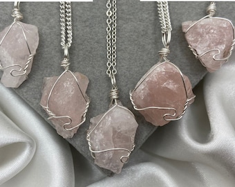 May include: Five silver necklaces with pink rose quartz pendants wrapped in silver wire. The necklaces are displayed on a white satin fabric background.