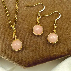 May include: A gold-coloured jewellery set comprising a necklace and earrings. The necklace has a fine chain with a pendant, and the earrings are drop style. Each piece features a round, pale pink gemstone, wire-wrapped in gold.