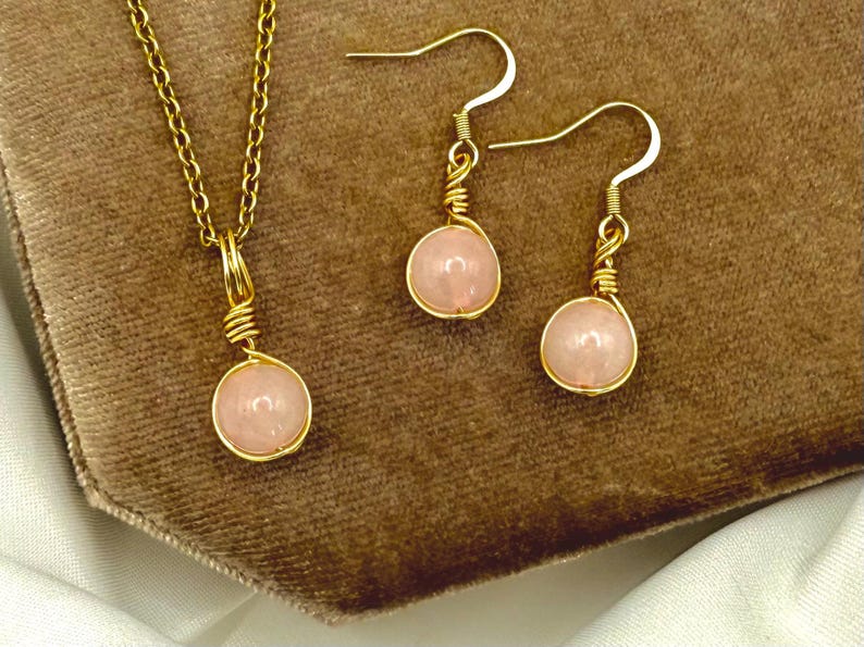 May include: A gold-coloured jewellery set comprising a necklace and earrings. The necklace has a fine chain with a pendant, and the earrings are drop style. Each piece features a round, pale pink gemstone, wire-wrapped in gold.