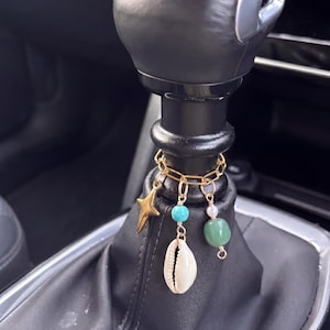 Può includere: Un accessorio a catena in metallo dorato avvolto attorno a un pomello del cambio nero di un'auto. Il ciondolo presenta una stella a quattro punte, una conchiglia di ciprea bianca con una perlina turchese e una pietra verde con una piccola perla.