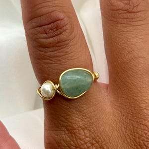 May include: A gold wire-wrapped ring with a light green, oval-shaped stone and a small, round white pearl. The ring is set against a soft, white background, highlighting the jewelry's design and colour.