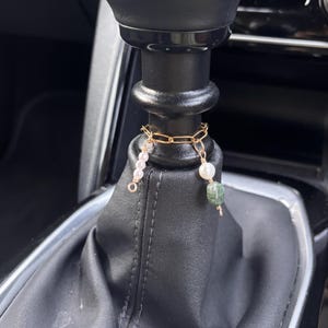 Op de afbeelding: Een goudkleurige ketting met bungelende bedels, waaronder parels en groene kralen, hangt aan de versnellingspook van een auto. De versnellingspookhoes is van zwart leer, met de binnenkant van de auto op de achtergrond.