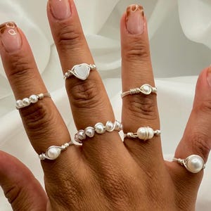 Puede incluir: Una mano muestra una colección de anillos hechos a mano con alambre. Los anillos presentan perlas y un dije en forma de corazón. Los anillos son de color plateado y varían en diseño, mostrando una estética delicada y elegante.