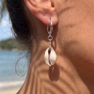 Op de afbeelding: Zilveren oorring met een kleine, natuurlijke schelpenhanger. De schelp is wit met een bruin interieur. De oorring wordt aan een oor gedragen, met een strandscène op de achtergrond.