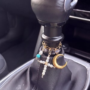 May include: A gold-coloured car gear shift charm featuring a cross, crescent moon, and chain design. The cross is decorated with white pearls, and a turquoise bead is also present. The charm hangs from a black gear shift.