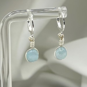May include: A pair of silver hoop earrings with light blue aquamarine stones hanging from them. The earrings are displayed on a clear acrylic stand.