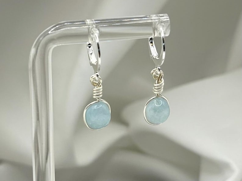 May include: A pair of silver hoop earrings with light blue aquamarine stones hanging from them. The earrings are displayed on a clear acrylic stand.