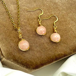 May include: A gold-coloured jewellery set comprising a necklace and earrings. Each piece features a pink gemstone pendant. The necklace has a fine chain, and the earrings have a hook design.