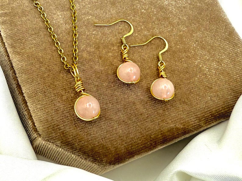 May include: A gold-coloured jewellery set comprising a necklace and earrings. Each piece features a pink gemstone pendant. The necklace has a fine chain, and the earrings have a hook design.