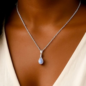 Pode incluir: Um colar de prata com um pingente em forma de gota, azul pálido. O pingente é enrolado em arame e suspenso por uma corrente delicada. O colar é mostrado em um fundo de tom de pele.