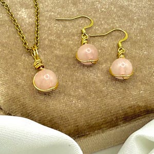 May include: A gold-tone necklace and earring set with pink gemstone beads. The necklace has a pendant with a round pink bead, and the earrings have matching beads. The beads are wrapped in gold wire.