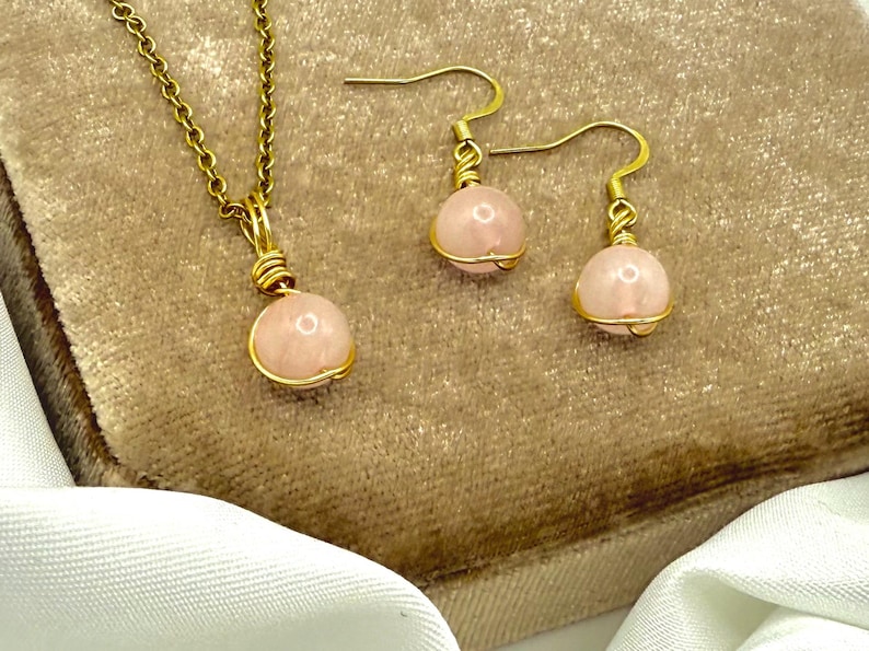 May include: A gold-tone necklace and earring set with pink gemstone beads. The necklace has a pendant with a round pink bead, and the earrings have matching beads. The beads are wrapped in gold wire.