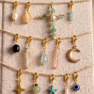 May include: A selection of gold-coloured charm pendants featuring diverse designs, such as a pearl, crystals, a cross, a crescent moon, a star, a turquoise starfish, and an evil eye. The charms are displayed on twine.