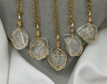 May include: Five gold necklaces with clear quartz crystal pendants wrapped in gold wire. The necklaces are displayed on a gray surface with white fabric.
